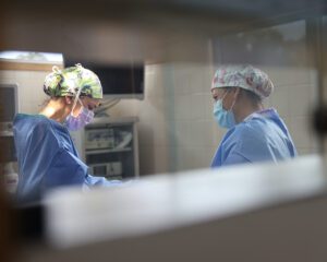 Two surgical team members in scrubs and masks during a procedure, viewed through an operating room window